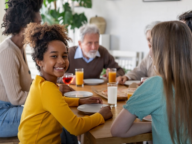 Menina sorrindo para a câmera enquanto está sentada à mesa de jantar com sua família.